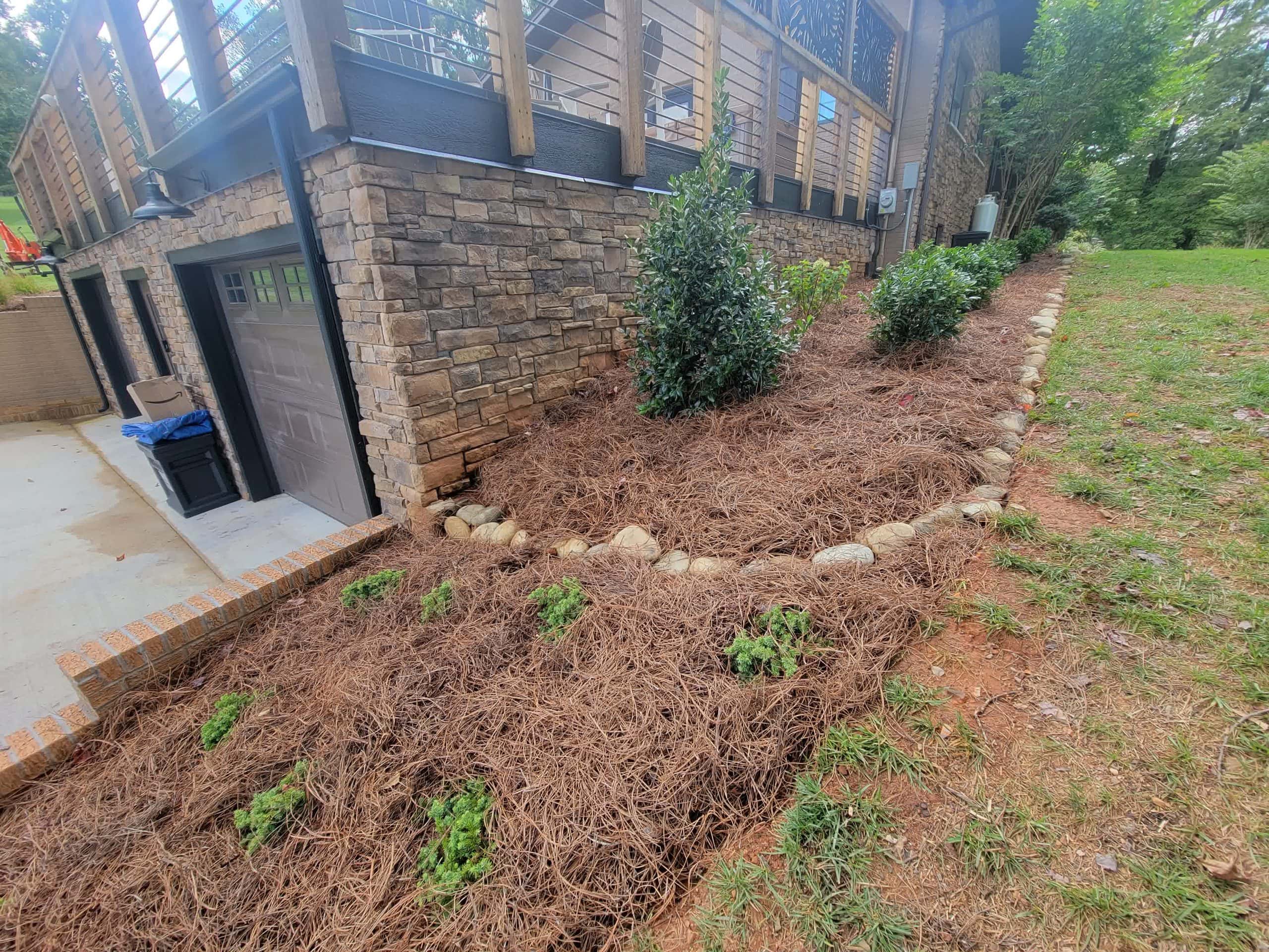 Sloped landscape with pine straw mulch, shrubs, and a stone border along a modern home with a raised deck and stone facade.