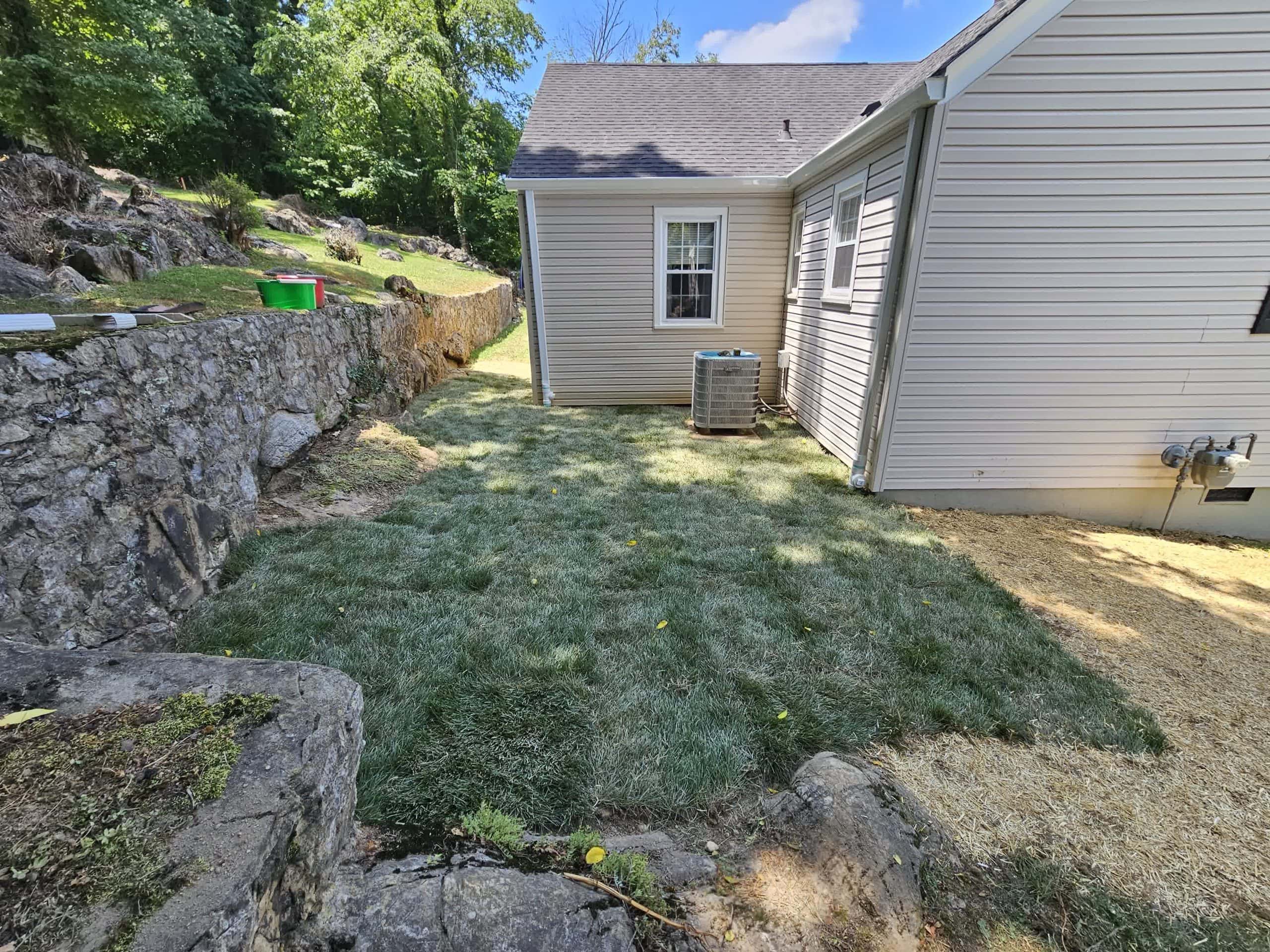 Backyard with freshly installed sod, a stone retaining wall, and an air conditioning unit beside a beige house.
