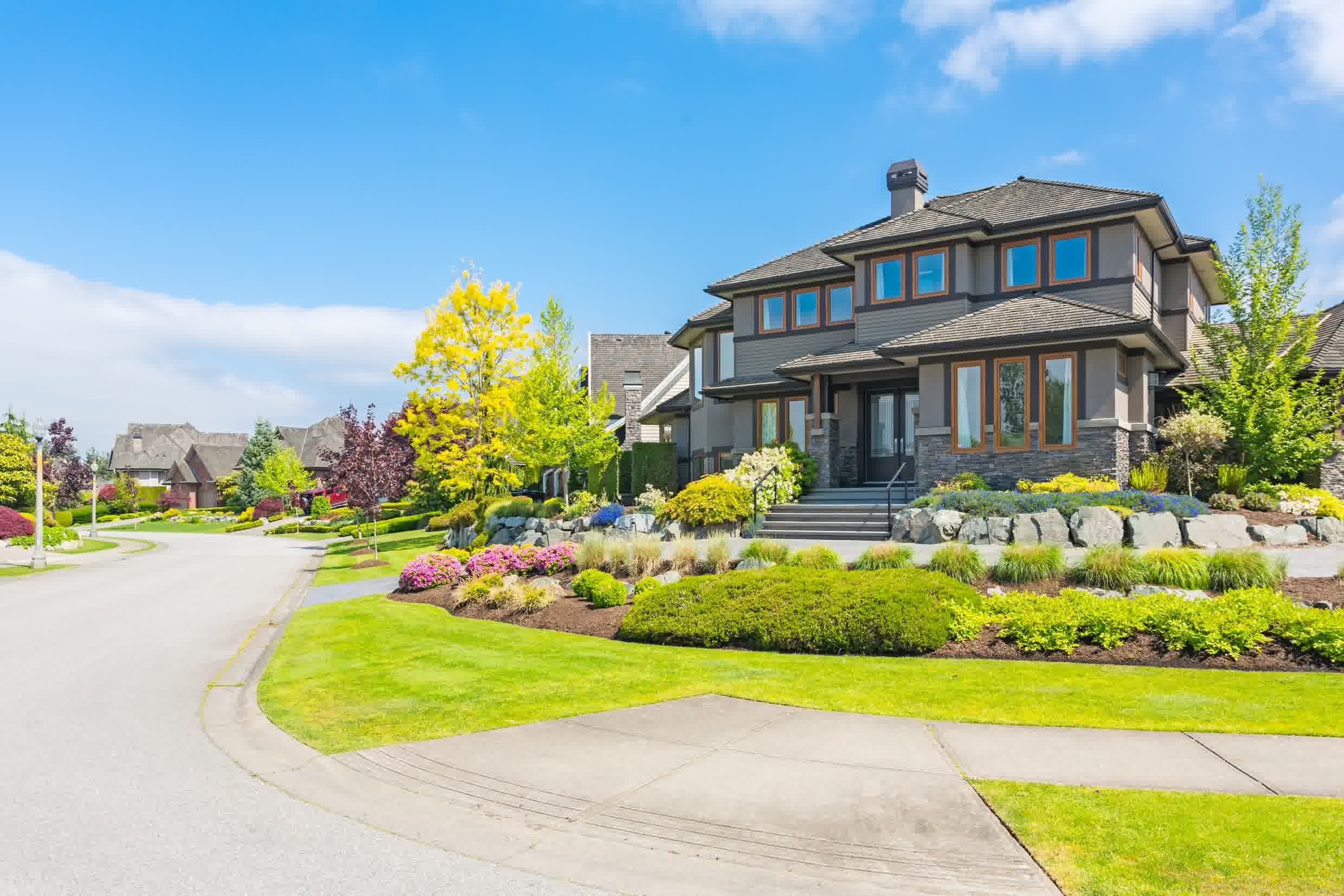Modern house with vibrant landscaping, colorful flower beds, and a lush front yard on a sunny day in a suburban neighborhood.