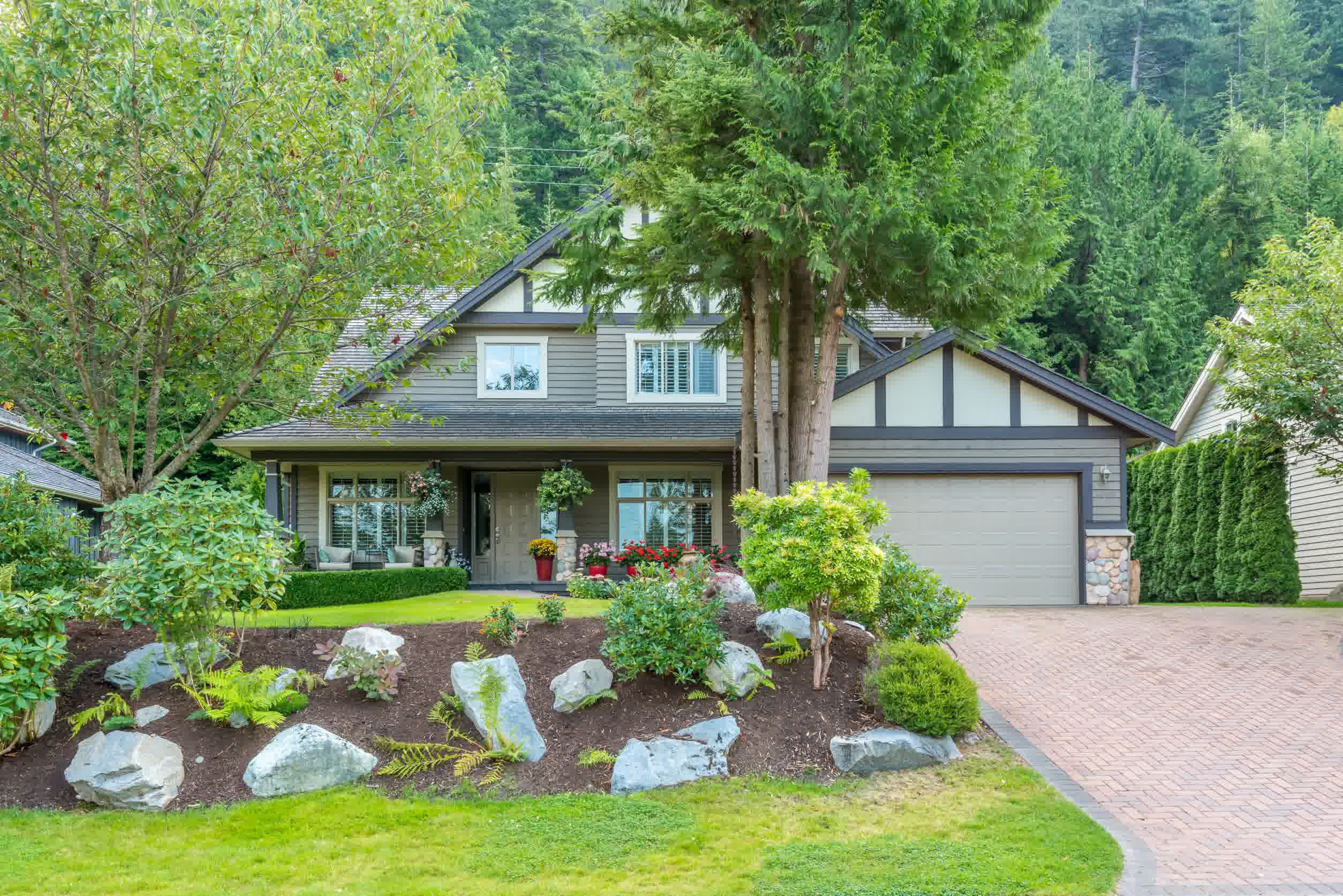 Charming house with a stone landscaping bed, lush plants, and a paved driveway framed by trees in a tranquil neighborhood.