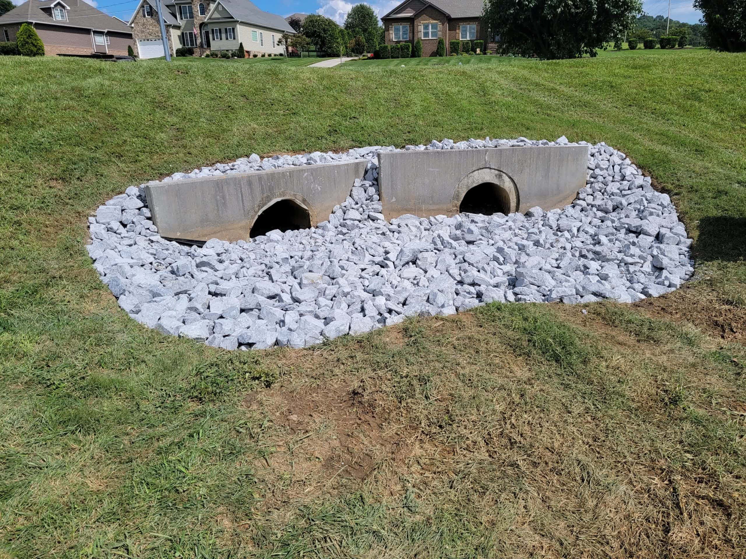 Dual concrete drainage culverts surrounded by white riprap stones on a sloped grassy yard to control erosion and improve water flow.