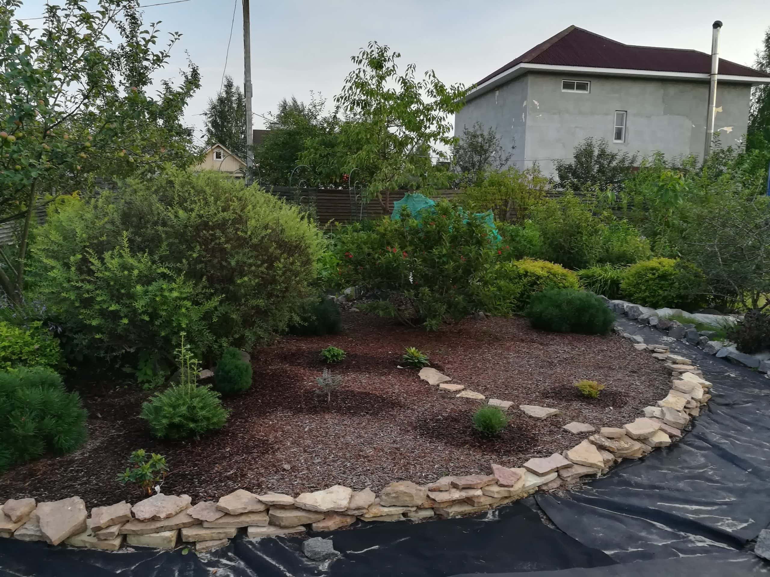 Rock-edged landscaping bed with mulch, shrubs, and young trees in a garden near a modern house.