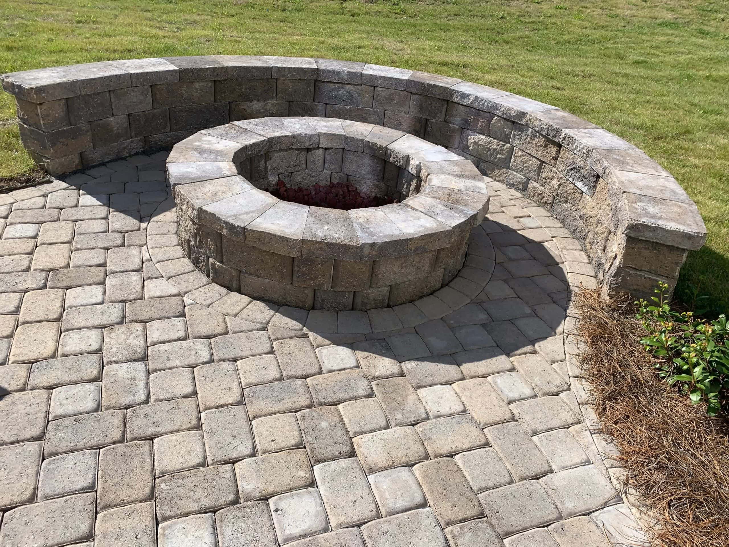 Circular stone fire pit with a built-in seating wall on a paver patio, surrounded by grass and minimal landscaping.