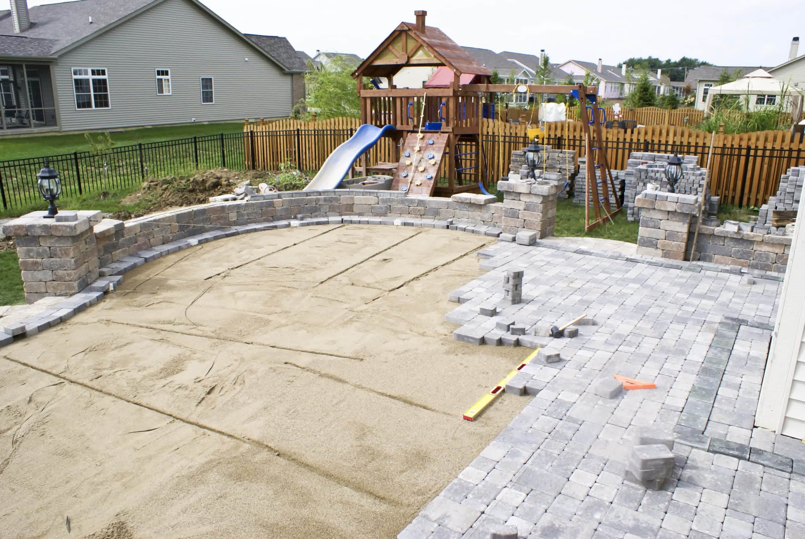 Large backyard paver patio under construction with retaining walls, level sand base, and a children’s playset in the background.
