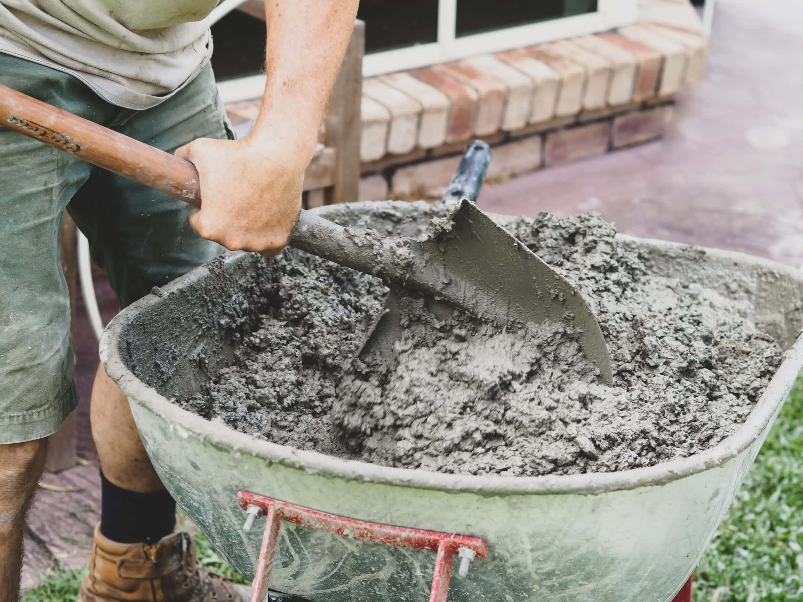 Construction worker mixing wet concrete in a wheelbarrow with a shovel, preparing material for a patio or masonry project.