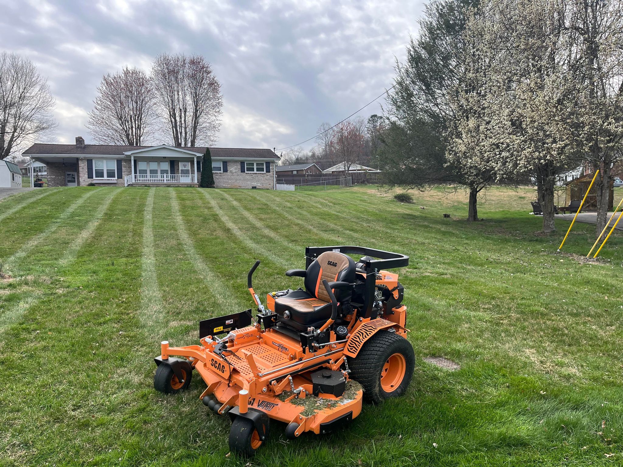 Bright orange SCAG zero-turn lawn mower parked on freshly striped lawn in front of a brick home with large front yard and blooming trees.